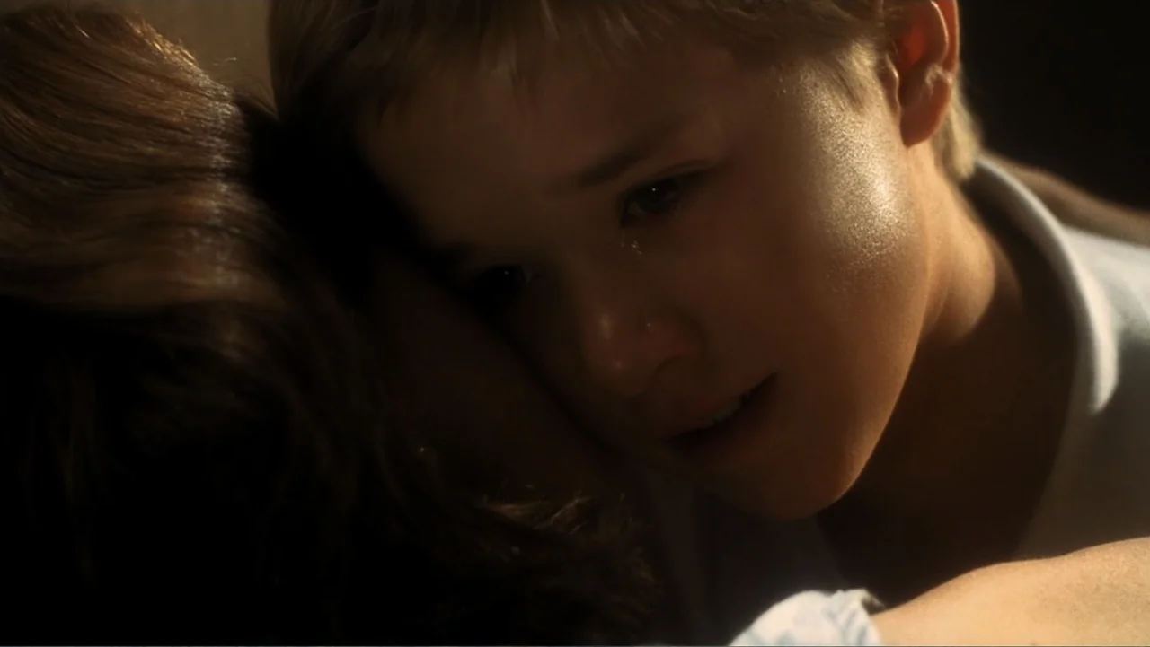 A.I. Artificial Intelligence (2001) movie still: closeup — A dark indoor space where a child rests against an adult figure. The boy leans his head…