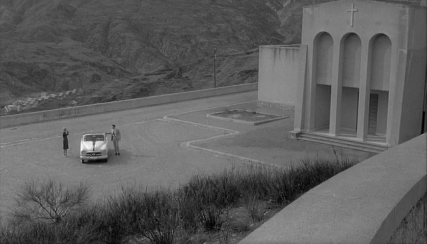 L’Avventura (1960) movie still: wide — A man and a woman stand by their parked car in a mountain parking area. A small modern church with three…