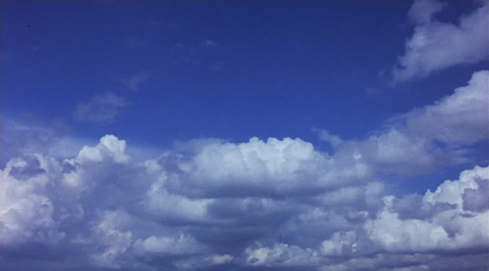 Groundhog Day (1993) movie still: establishing — A vast blue sky is filled with large, fluffy white cumulus clouds. The clouds stretch across the…