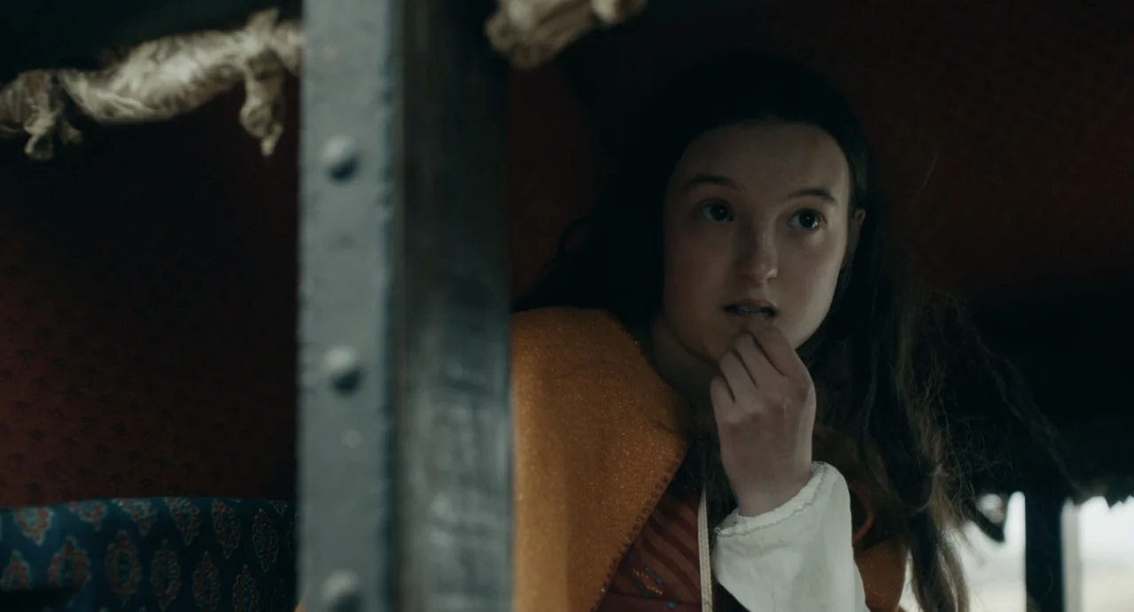 Catherine Called Birdy (2022) movie still: closeup — A dimly lit wooden interior of a horse-drawn carriage. A young girl sits inside looking outward…