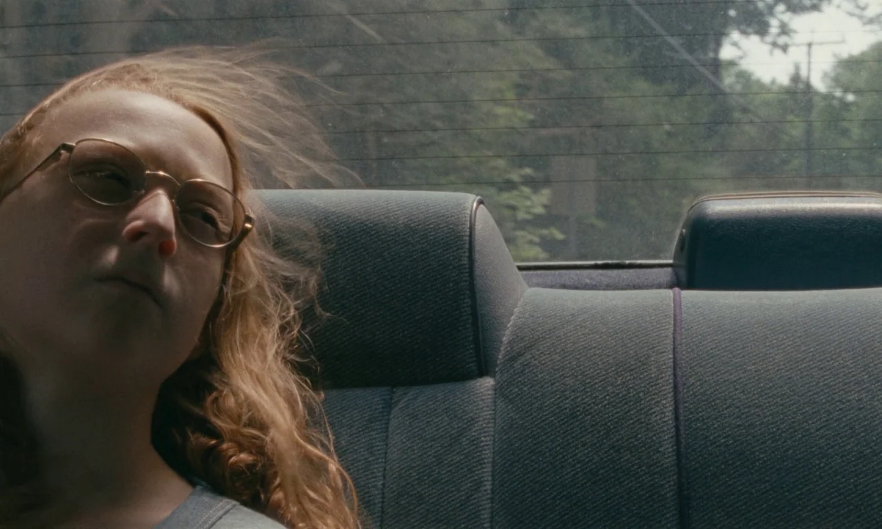 Janet Planet (2023) movie still: closeup — A person sits in the back of a moving car with blurred greenery outside. Wind blows through her hair as…
