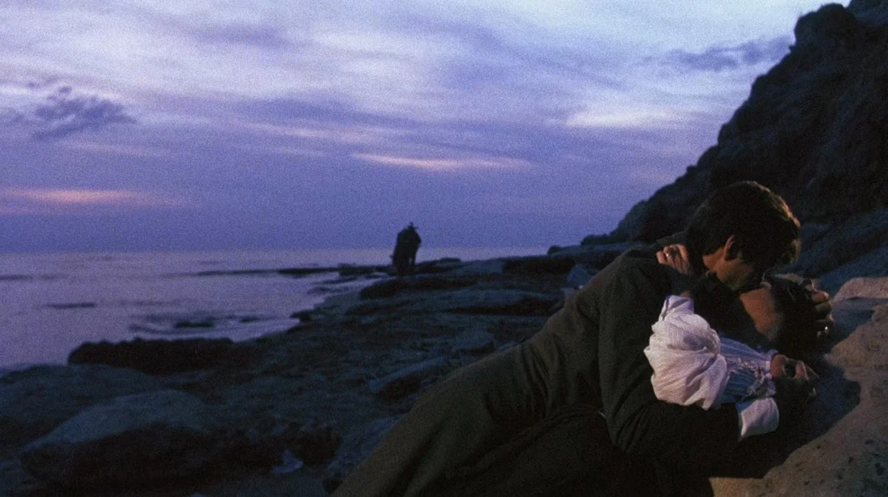 The Whip and The Body (1963) movie still: medium — A rocky coastal shoreline under a deep purple dusk sky with silhouettes of cliffs. A man and a…
