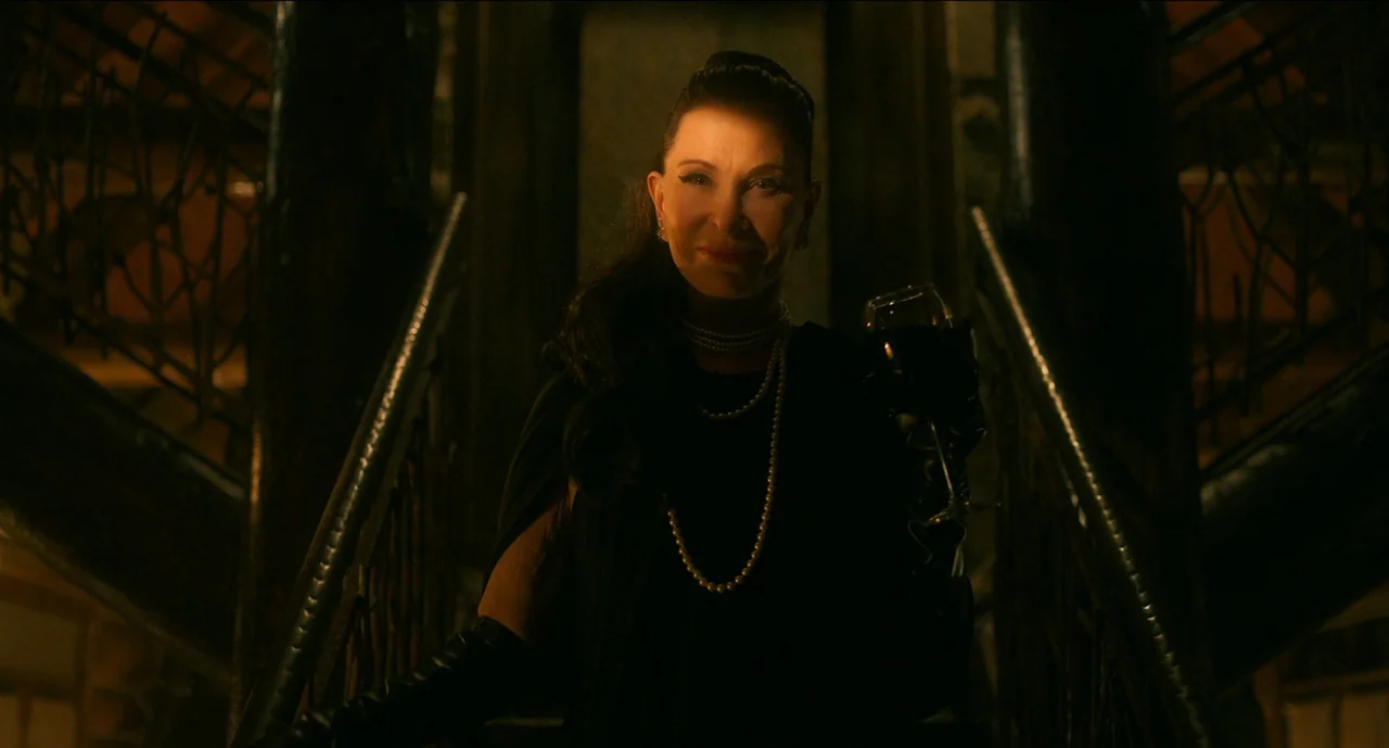Trim Season (2023) movie still: medium — A woman stands on a dimly lit staircase flanked by metal railings. She holds a wine glass in one hand while…