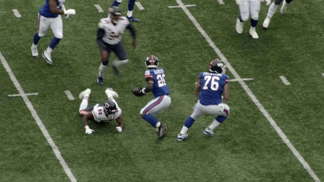 SAQUON (2025) movie still: wide — An outdoor professional sports field with white yard lines and artificial green turf. A player runs with the…