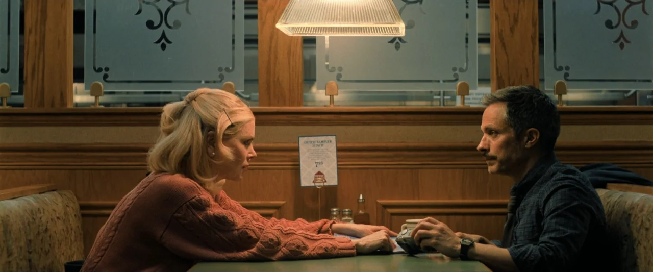 Holland (2025) movie still: medium — A diner booth with wood-paneled walls features decorative etched glass partitions above the seats. A man and a…
