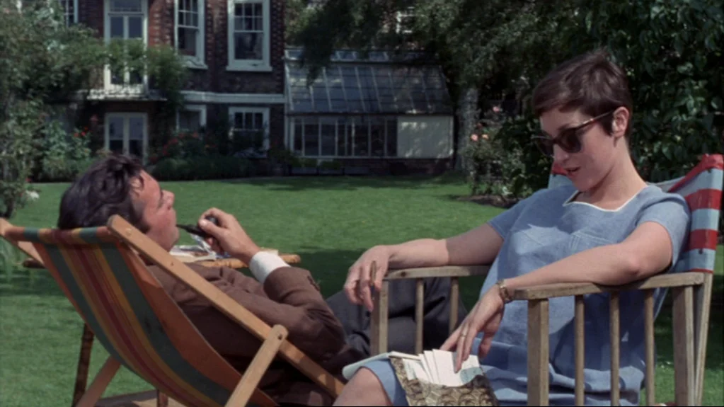 Accident (1967) movie still: medium — A manicured lawn features a large brick residence with a greenhouse in the background. A man and a woman sit…