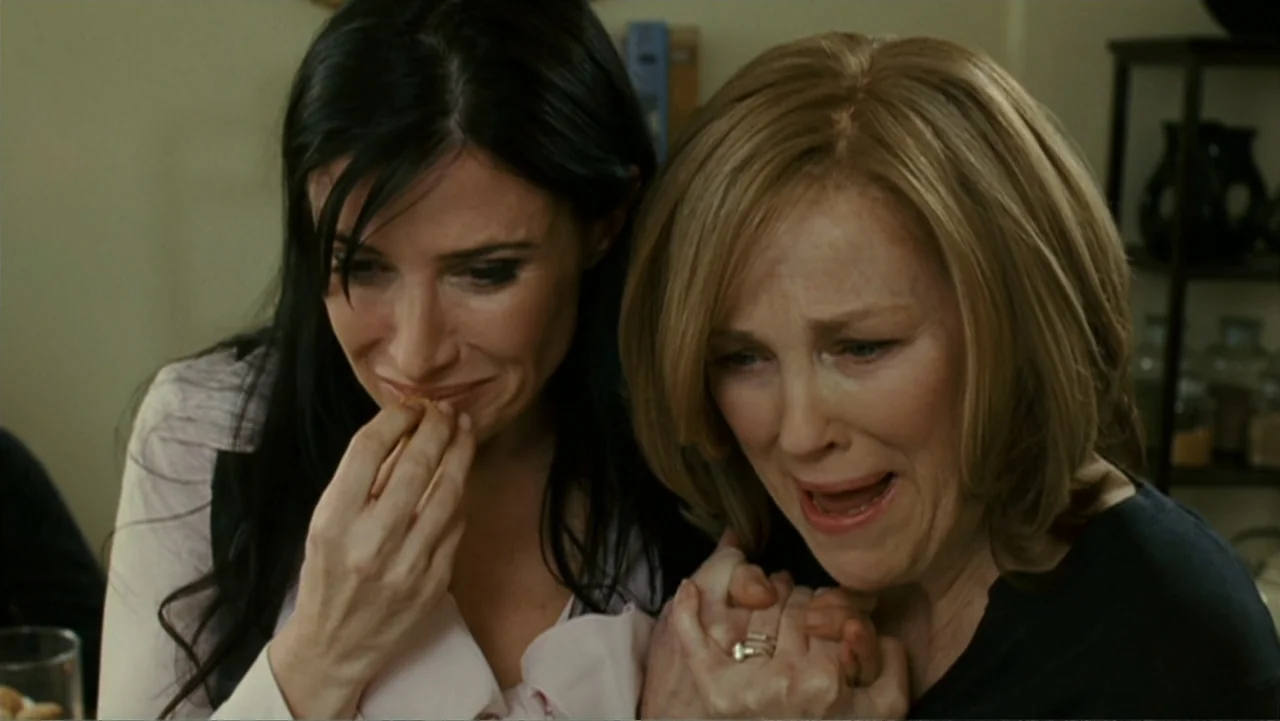 Penelope (2007) movie still: medium — A quiet indoor space with soft beige walls and simple decor. Two women sit side by side holding hands while…