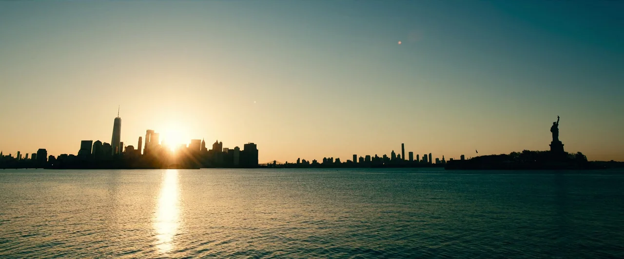 Leave The World Behind (2023) movie still: establishing — A wide view of the New York City skyline at sunset across the harbor. The sun sits low on…