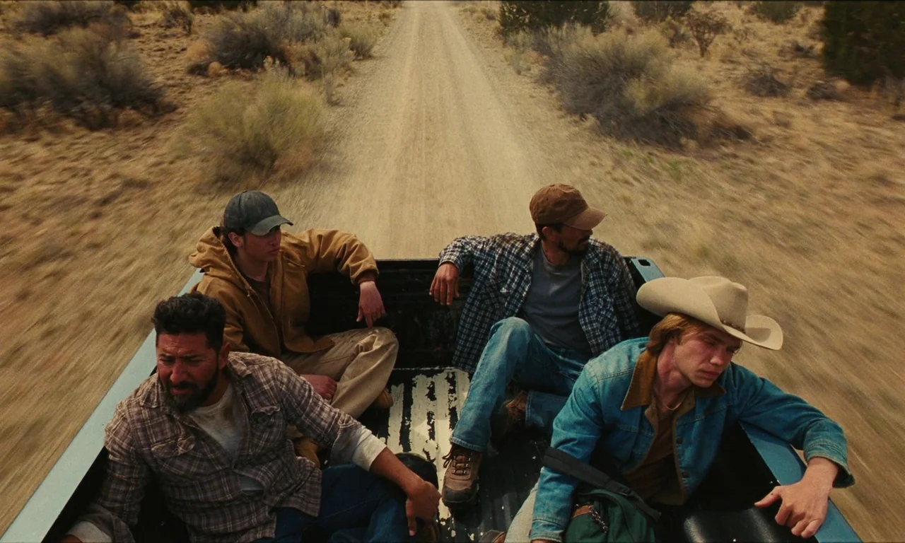 National Anthem (2023) movie still: wide — A group of four men ride in the back of a moving pickup truck. They are traveling along a dusty dirt…