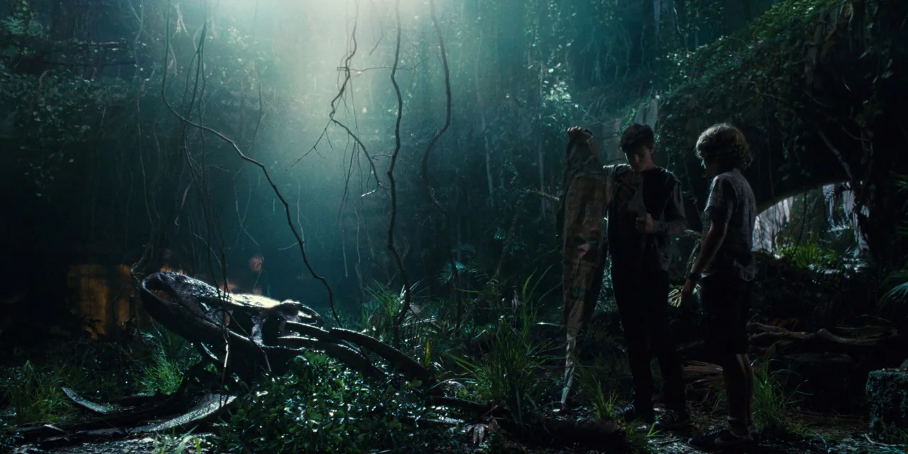 Jurassic World (2015) movie still: wide — A dense, overgrown jungle clearing features a large, weathered animal skull on the ground. Two teenage…