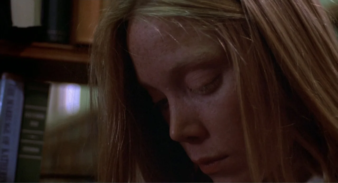 Carrie (1976) movie still: closeup — A quiet library corner contains rows of books on dark wooden shelves. A young woman with blonde hair bows her…