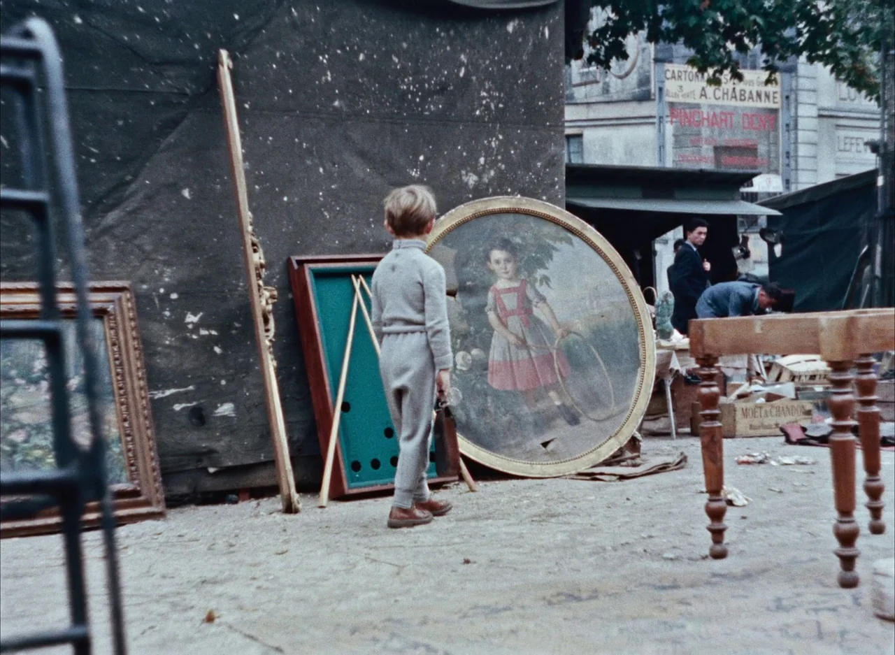 The Red Balloon (1956) movie still: fullbody — An outdoor flea market contains various antique objects, including furniture and framed art pieces. A…