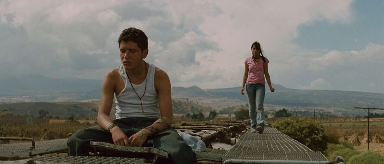Sin Nombre (2009) movie still: fullbody — The roof of a freight train travels through a vast rural landscape with distant hills. A man sits facing…