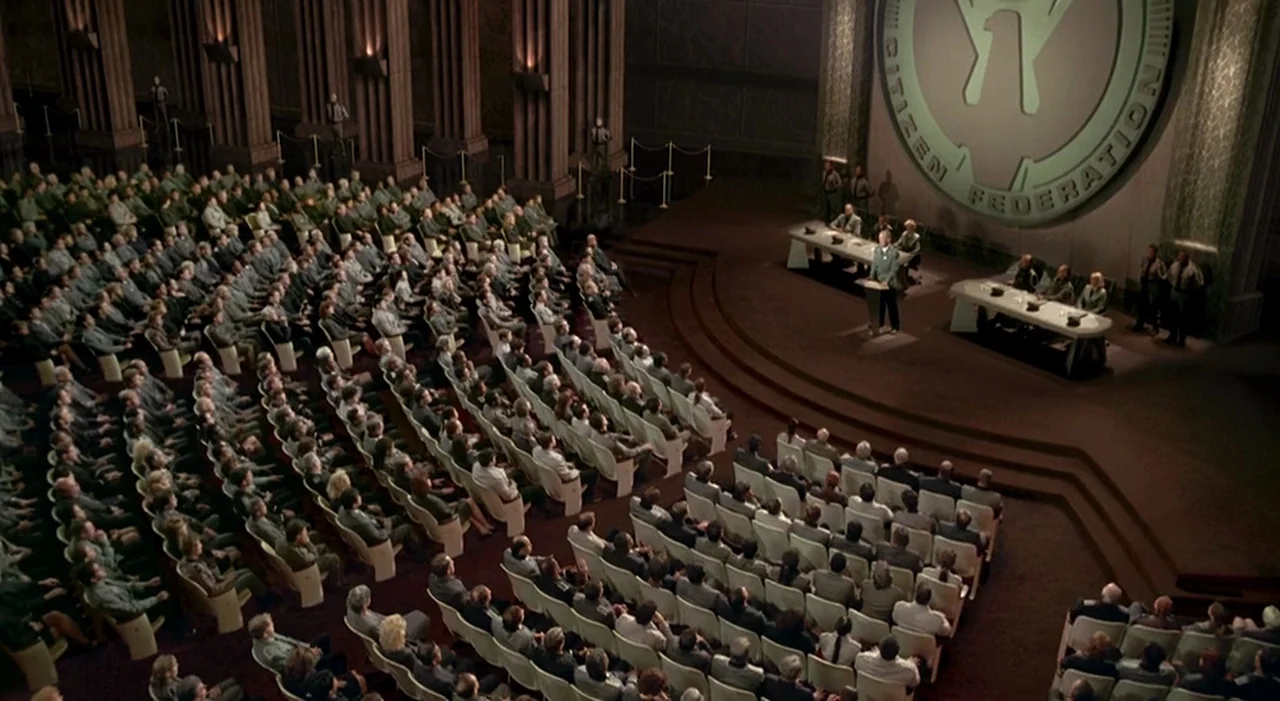 Starship Troopers (1997) movie still: wide — A vast, tiered auditorium features a large wall emblem for the Citizen Federation above. A single…