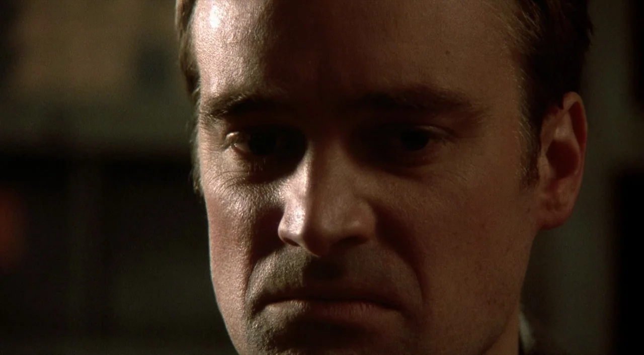 Cube (1997) movie still: closeup — A close-up view of a man in a dark, dimly lit indoor setting. The man stares forward with a serious facial…