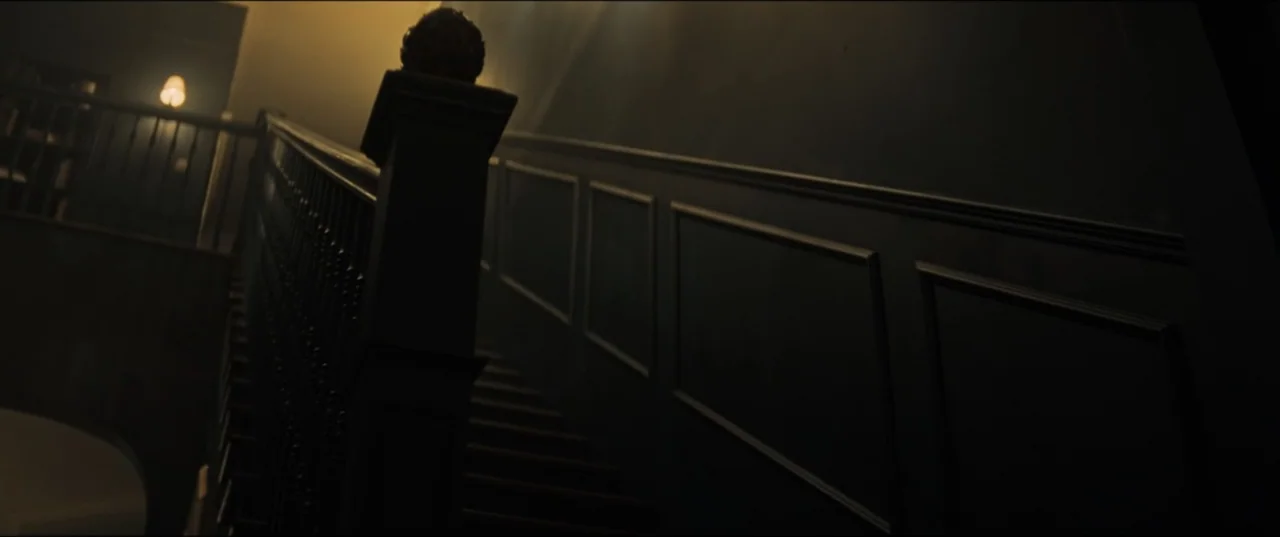 Vicious (2025) movie still: establishing — A dark wooden staircase features a prominent carved newel post and panelled wall. A lone warm lamp glows…