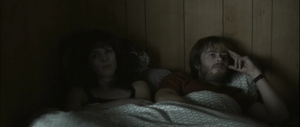 One Hundred Mornings (2009) movie still: medium — A dimly lit room with wood-paneled walls and patterned bedding. A woman and a man lie together in…