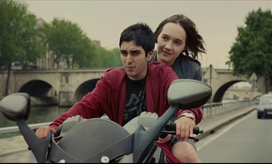Hadewijch (2009) movie still: medium — A pair of young people travel on a motorcycle along a paved riverside road. Stone arches of a bridge span the…