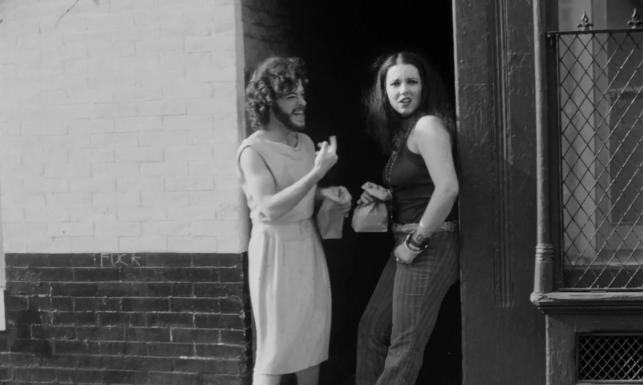 Multiple Maniacs (1970) movie still: fullbody — A brick building entrance with a decorative metal grate on the side window. Two people stand in the…