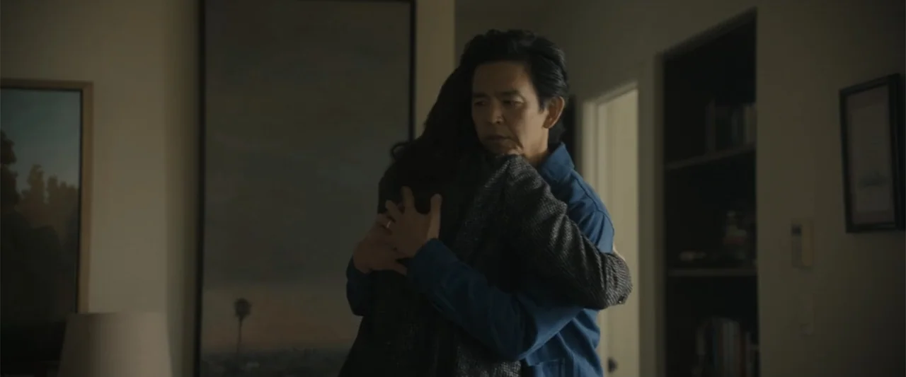 Afraid (2024) movie still: medium — An interior living room with muted beige walls and framed artwork on the wall. A man embraces an individual who…