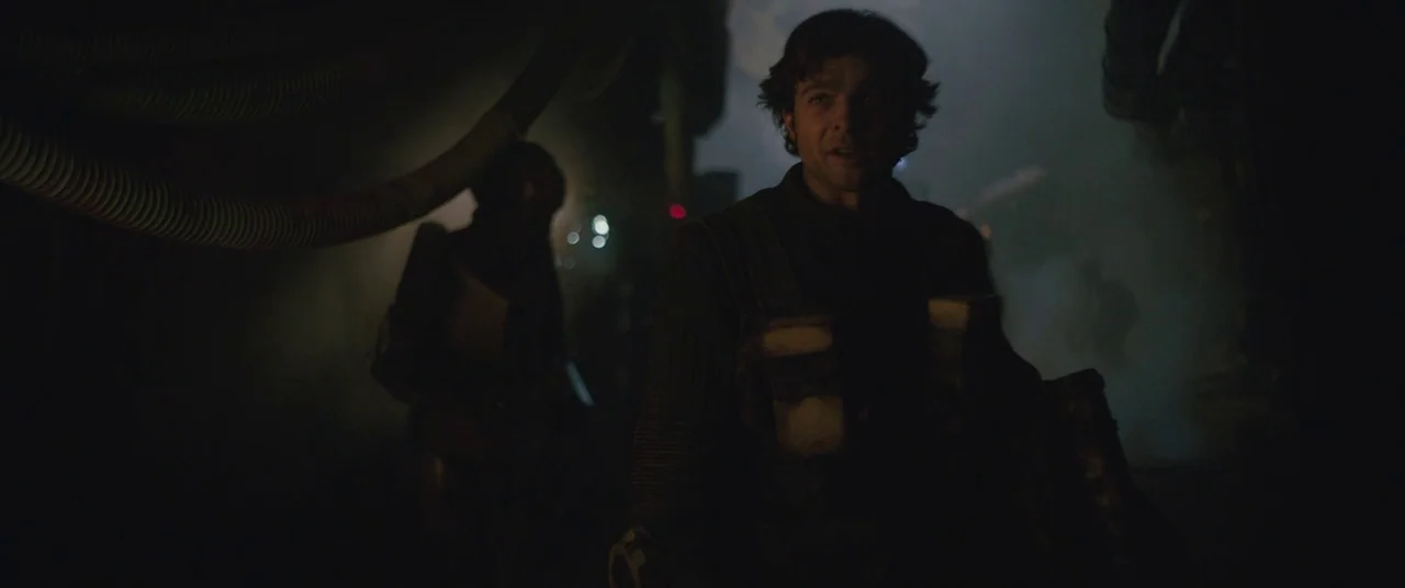 Solo: A Star Wars Story (2018) movie still: medium — A dimly lit, narrow industrial passage contains thick pipes and heavy shadows. A man with short…