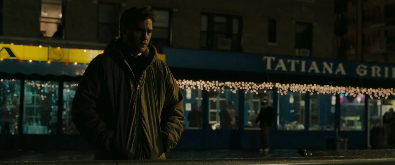 Two Lovers (2008) movie still: medium — A man stands on a dark sidewalk in front of a blue-painted restaurant facade. Strings of lights glow above…