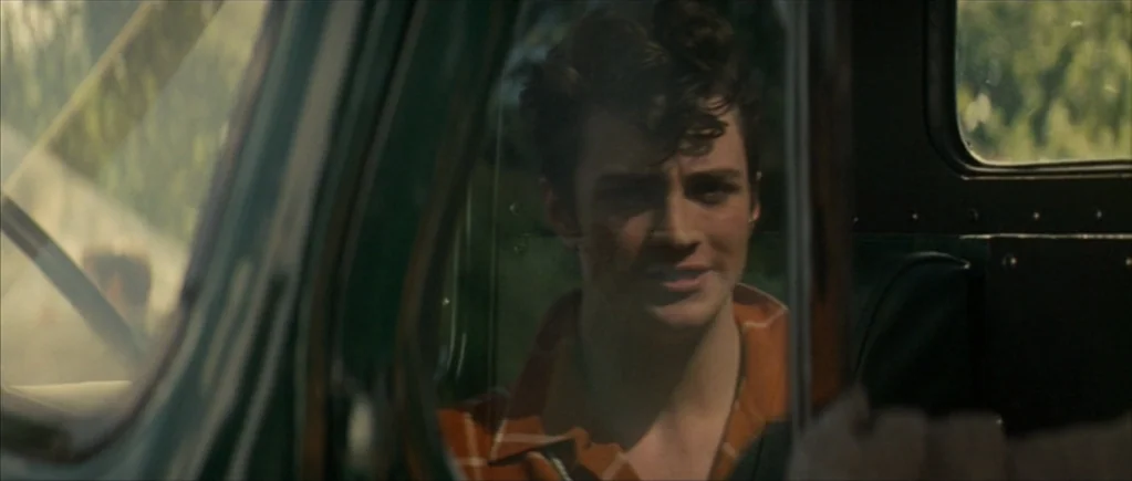 Nowhere Boy movie still: closeup — A man sits inside the cabin of a vintage truck, viewed through a side window. Greenery is visible in the blurry…