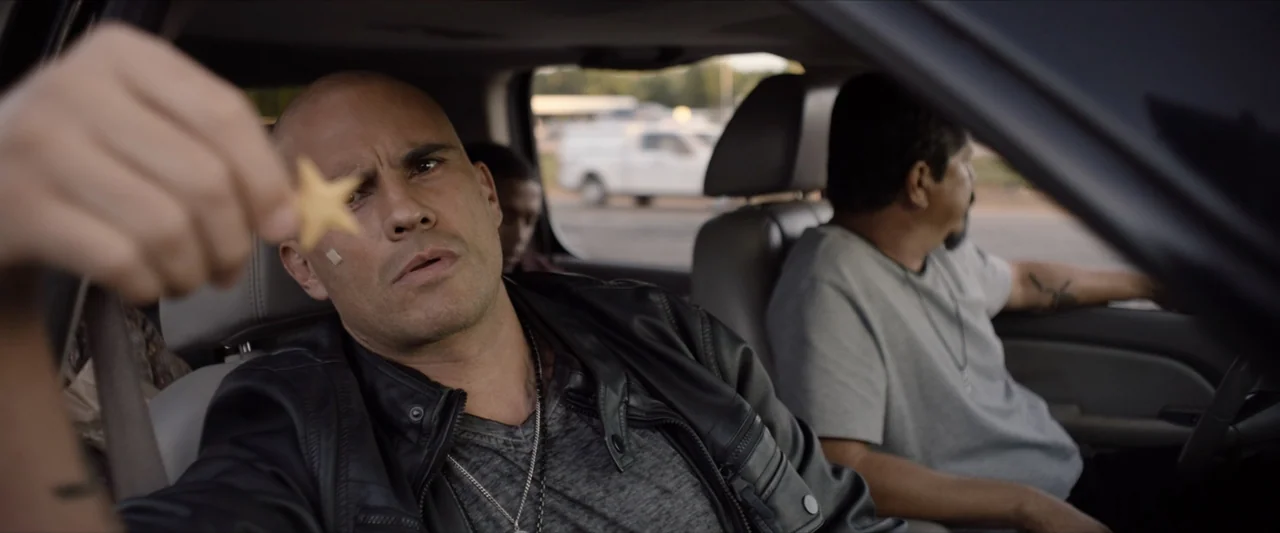 The Marksman (2021) movie still: medium — The interior of a parked car shows two men seated in the front row. The driver looks ahead while the…
