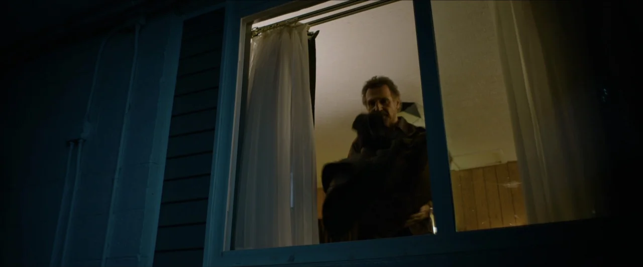 The Marksman (2021) movie still: medium — A dark outdoor view looks through a window into a dimly lit motel room. An older man stands inside the…