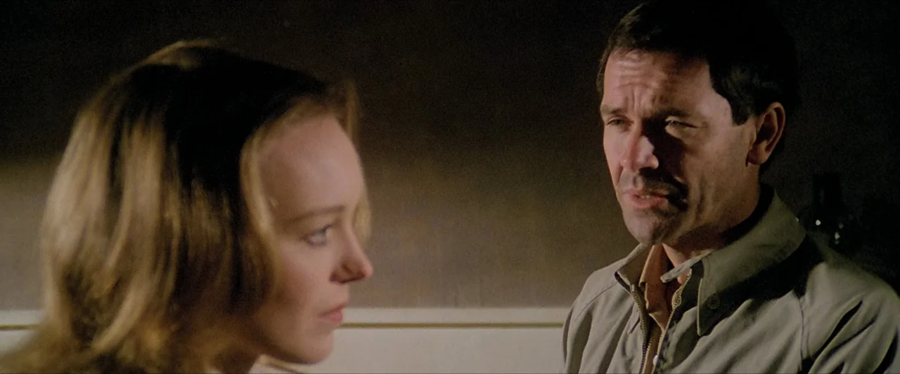 The Beyond (1981) movie still: medium — Two individuals stand facing each other in a dimly lit, nondescript room. The man in the foreground speaks…