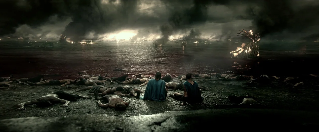 300: Rise of an Empire (2014) movie still: wide — A dark, bloody shoreline littered with fallen soldiers under a smoke-filled, stormy sky. Two men…