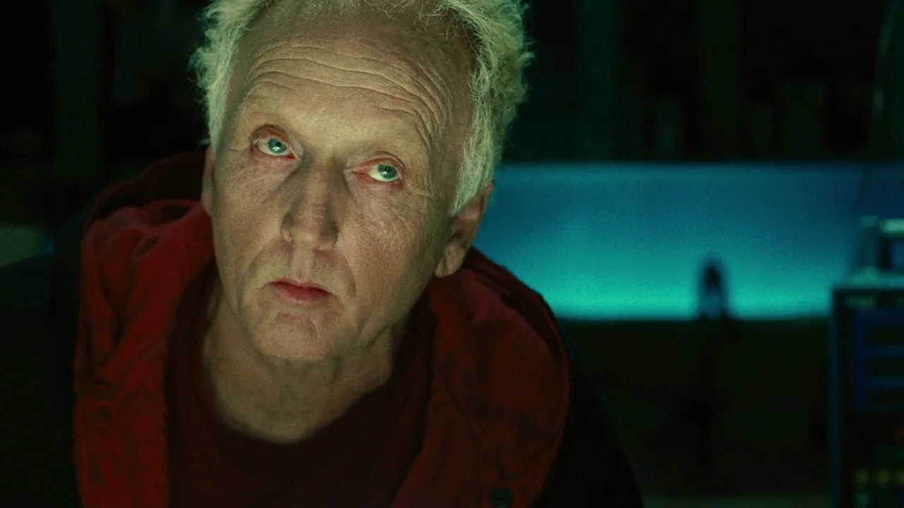 Saw II movie still: closeup — A dimly lit room bathed in cool green tones is the backdrop for the subject. A man with thinning hair stares forward…