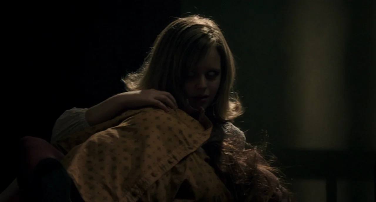 Ouija: Origin of Evil (2016) movie still: medium — A dark, dimly lit room contains a young woman with pale eyes. The person stares directly at the…