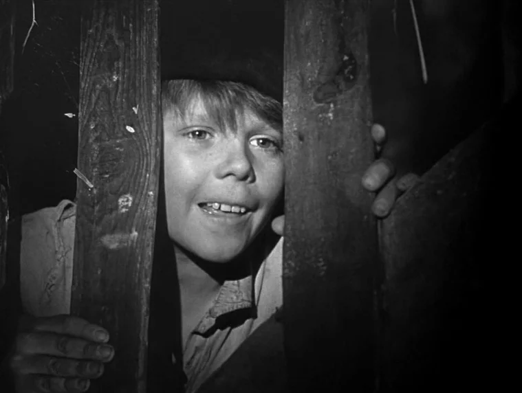 Forbidden Games (1952) movie still: closeup — A young boy peers between the vertical wooden boards of a dark fence. The illumination focuses on his…