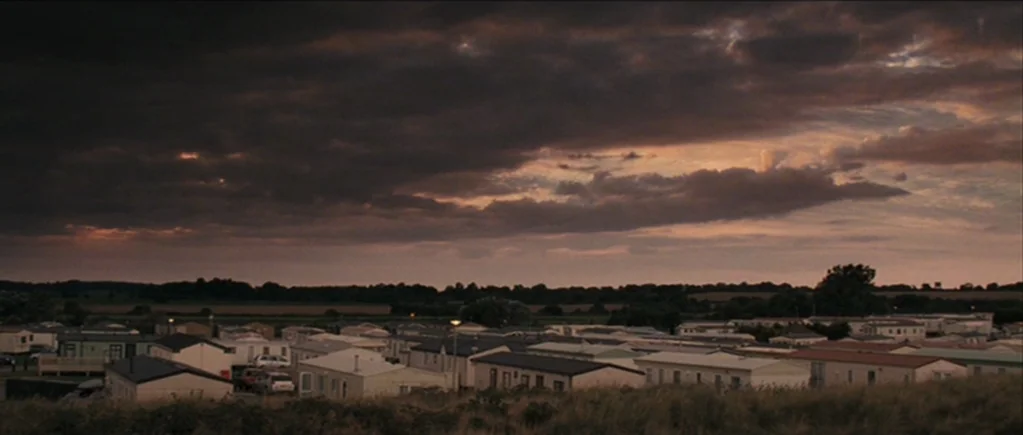The Scouting Book for Boys (2009) movie still: establishing — A dense collection of mobile homes sits beneath a vast, cloud-covered dusk sky. The…