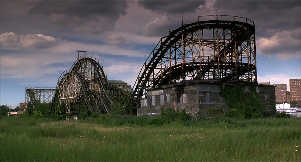He Got Game (1998) movie still: establishing — An abandoned wooden roller coaster structure stands overgrown with vegetation in a vacant field. Dark…
