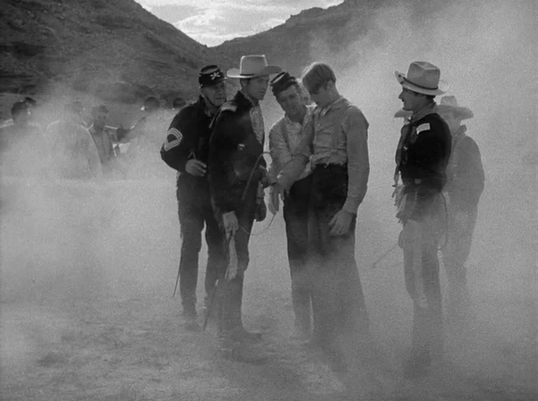 Rio Grande (1950) movie still: fullbody — A group of men in 19th-century western clothing stands in a thick dust cloud. Mountainous desert terrain…