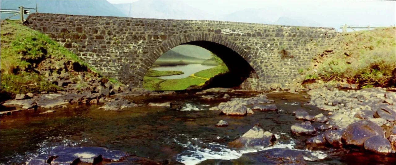 Breaking the Waves (1996) movie still: wide — A stone arch bridge spans a shallow river with exposed rocks in the foreground. Grassy slopes rise up…