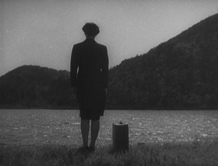 No Regrets for Our Youth (1946) movie still: fullbody — A woman stands on a grassy bank overlooking a calm body of water. She stands next to a small…