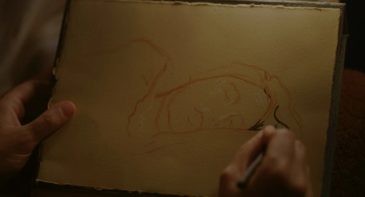 Portrait of a Lady on Fire movie still: closeup — A close-up view shows a hand sketching a portrait onto tan textured drawing paper. The person uses…