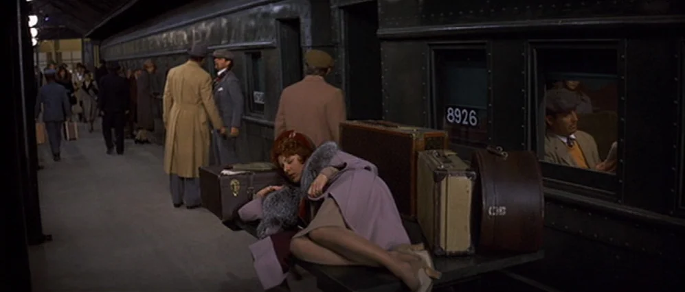 The Fortune (1975) movie still: wide — A bustling 1930s train platform with passengers standing near dark green locomotive carriages. A woman in a…