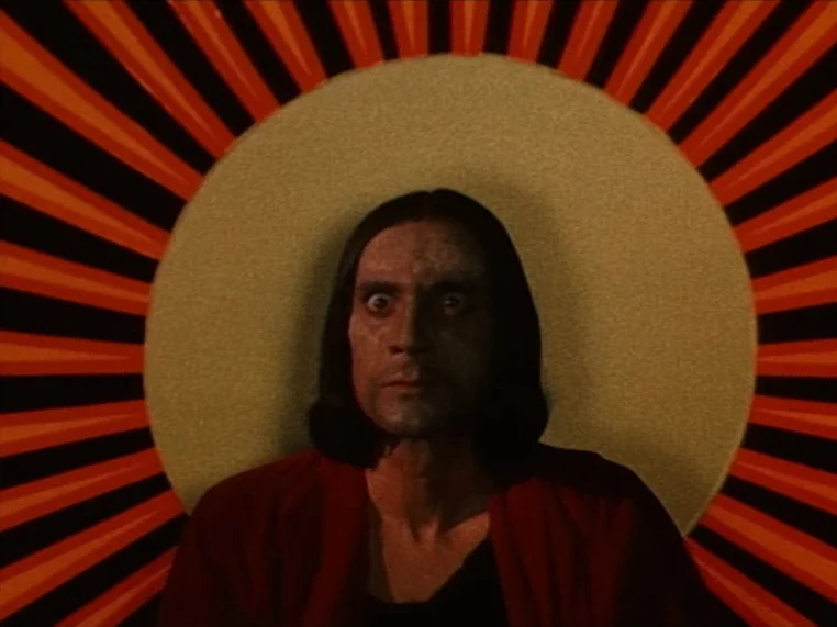 Salome (1972) movie still: closeup — A man stands in front of a backdrop featuring a giant sun graphic. Red and black sunrays radiate outward from…