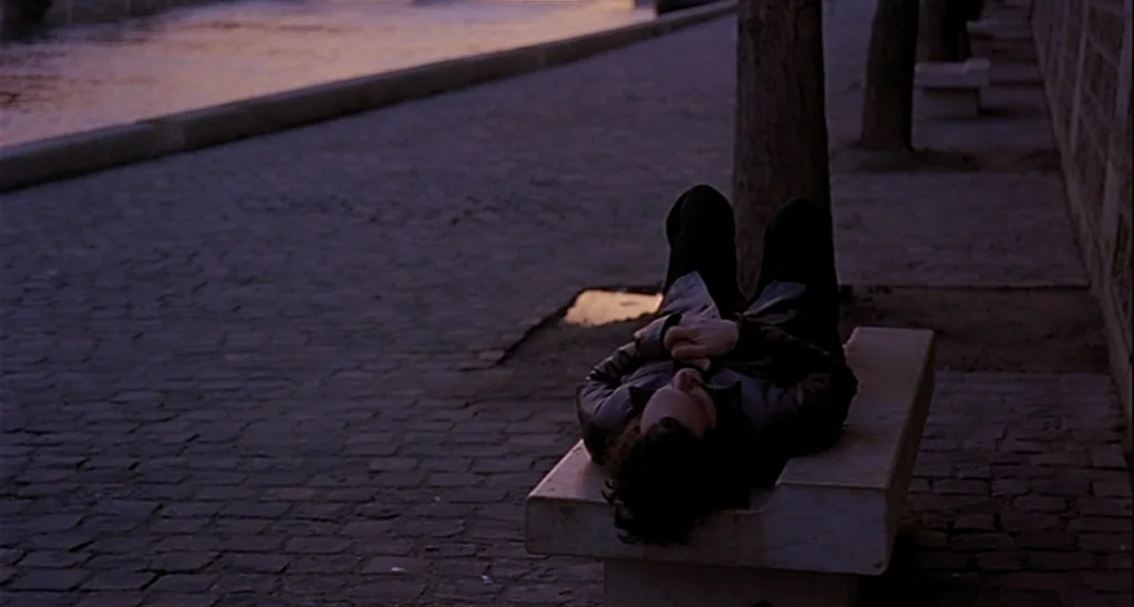 Trouble Every Day (2001) movie still: wide — A stone embankment beside a river features a smooth stone bench. A man lies on his back with his knees…