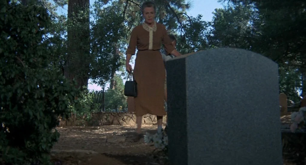 The Other (1972) movie still: fullbody — A quiet cemetery with mature trees and a prominent grey stone monument in foreground. A woman stands in…