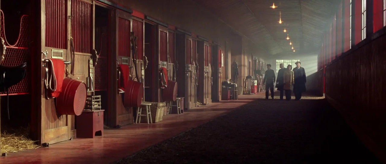 Seabiscuit (2003) movie still: wide — A long horse stable aisle features individual wooden stalls with red painted doors and hay. Four people walk…