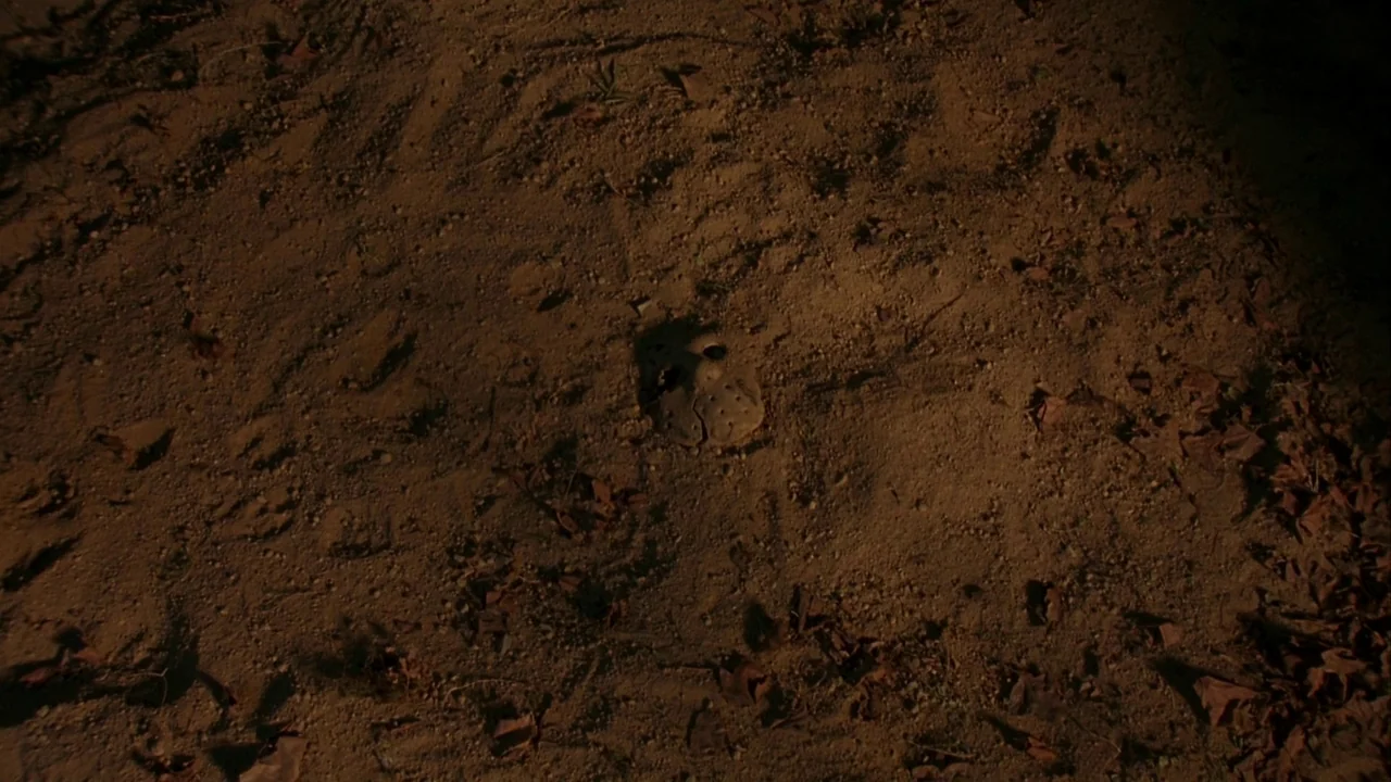 Jason Goes To Hell (1993) movie still: closeup — An abandoned hockey mask rests face up in the center of a dirt path. Dry leaves and coarse soil…