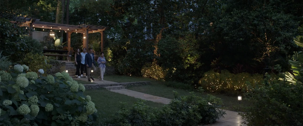 Father of the Bride (1991) movie still: wide — A residential backyard at night features a stone patio and wooden pergola overhead. Four people walk…