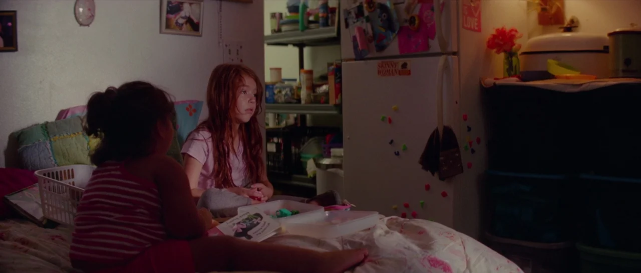The Florida Project (2017) movie still: medium — A dimly lit bedroom cluttered with personal items including photos, toys, and a refrigerator. Two…