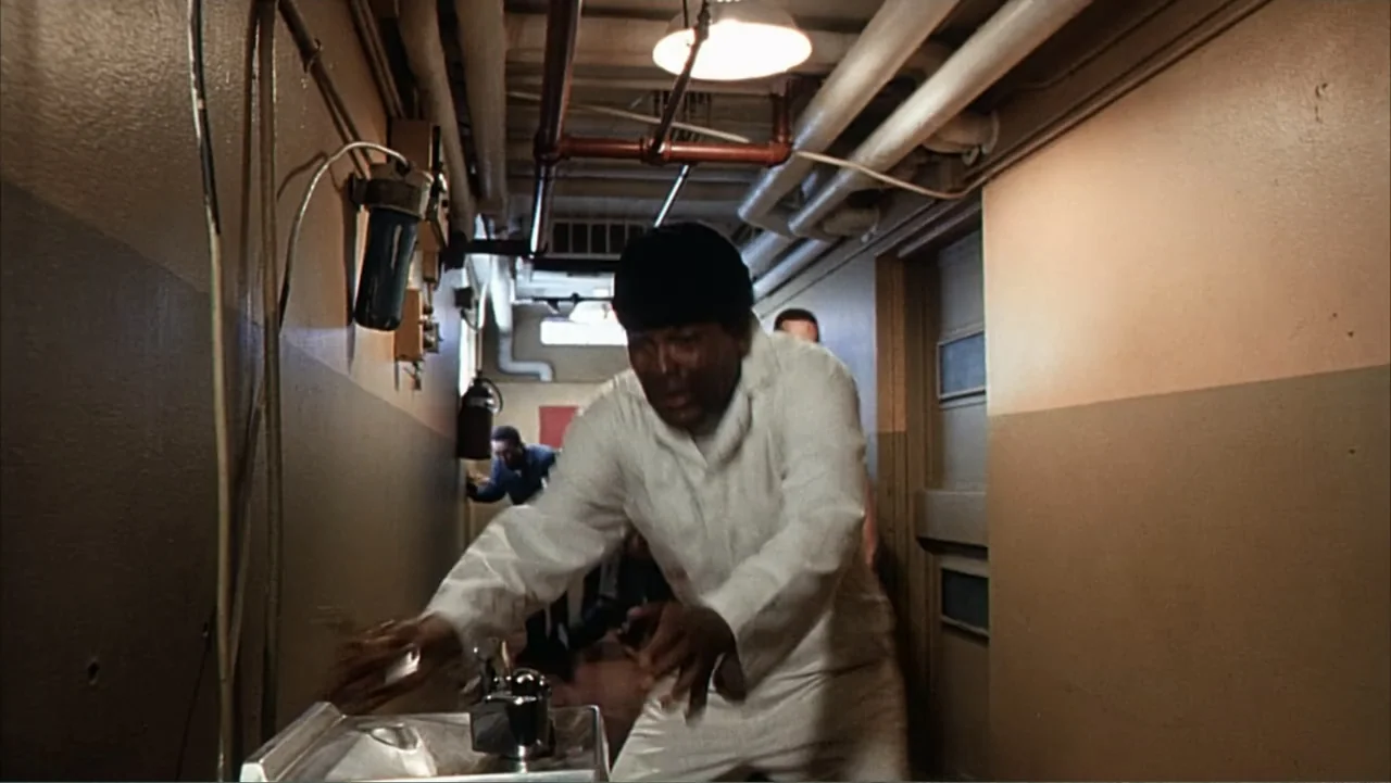 Against the Wall (1994) movie still: medium — A narrow institutional hallway has tan walls and exposed pipes on the ceiling. A man in white medical…