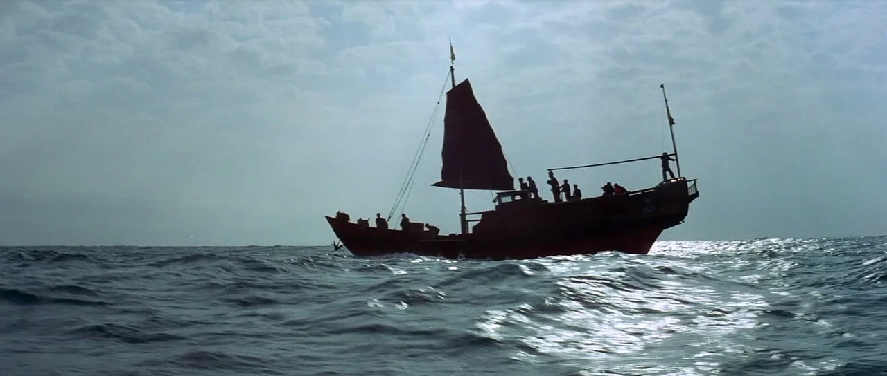 Enter The Dragon (1973) movie still: wide — A wooden junk boat with a dark sail travels across choppy open ocean water. Several figures are…